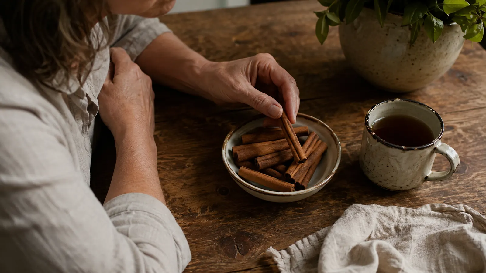 A warm kitchen counter with a small ceramic dish piled with whole cinnamon quills, a mug of tea, and a cloth napkin in soft morning light